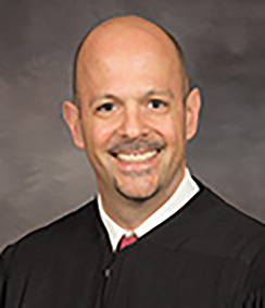 Judge Sam Salario headshot a Caucasian man whom is smiling, he has brown eyes in a black judges' robe with a white shirt and blue striped tie