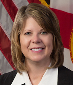 Judge Susan Kelsey headshot of a middle aged smiling Caucasian woman in a judges' robe with a white collared shirt. There is an American flag behind her.