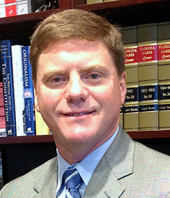 Judge Tim Osterhaus headshot of a middle aged Caucasian man with light brown hair. He is wearing a light grey suit, light colored shirt and blue striped tie. There is a bookshelf with large green books in the background.