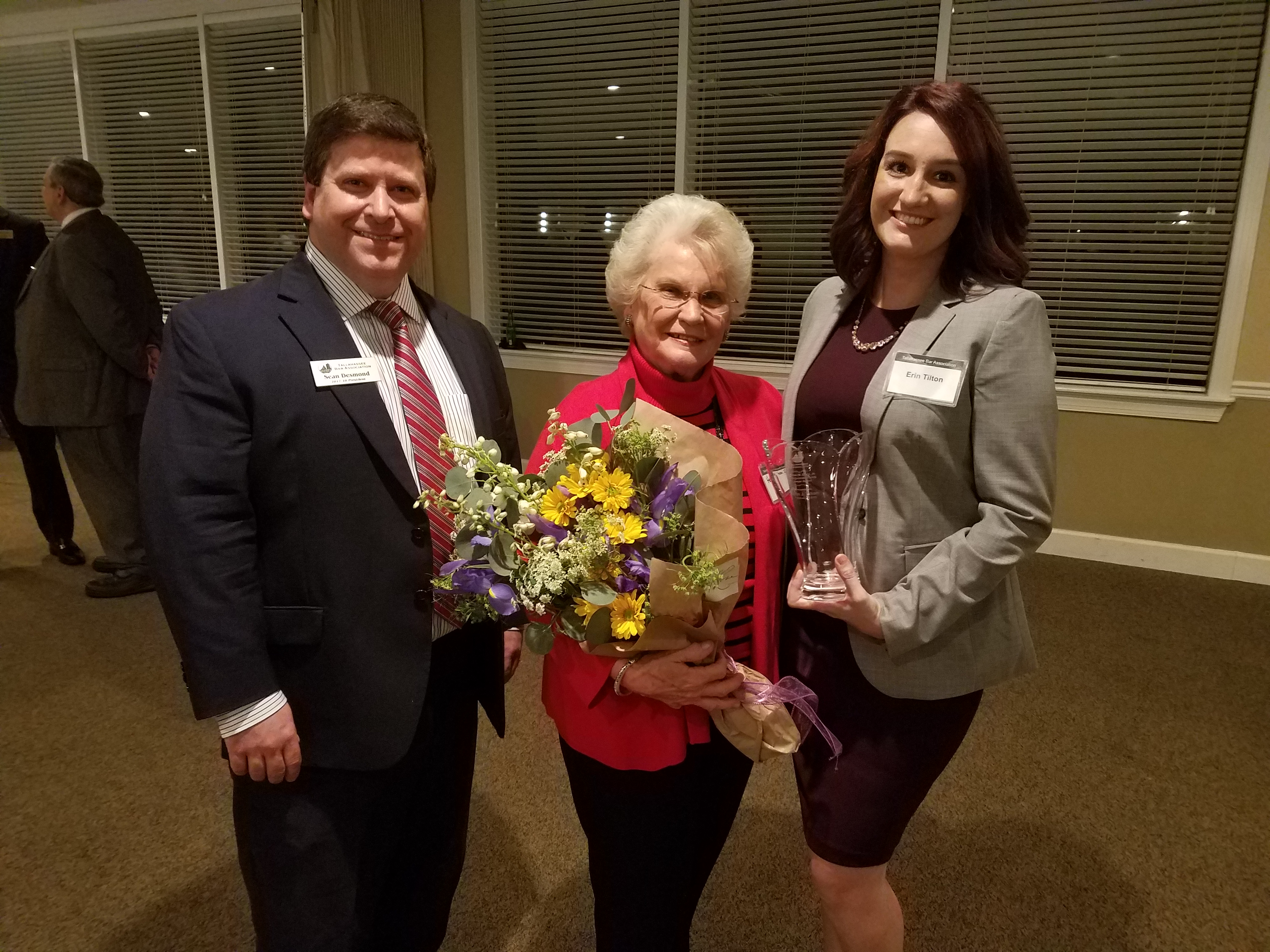 Sean Desmond a middle aged smiling Caucasian man in a black suit with brown hair and a red tie, Helen an older Caucasian woman in a red sweater holding a bouquet of flowers, Erin Tilton a young Caucasian woman that is smiling she is dressed in a grey blazer and it holding an award vase