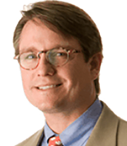 Curry Pajcic headshot of a cheerful Causasian man who is smiling. He has brown glasses and neatly combed brown hair. He is wearing a tan suit with blue shirt and a reddish orange tie.