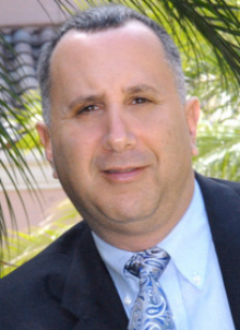 Joseph E Ankus headshot of a Caucasian man in a blue suit with a blue tie, there are palm trees in the background.
