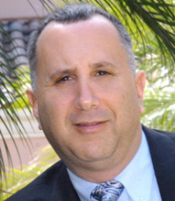 Joseph E Ankus headshot of a Caucasian man in a blue suit with a blue tie, there are palm trees in the background.