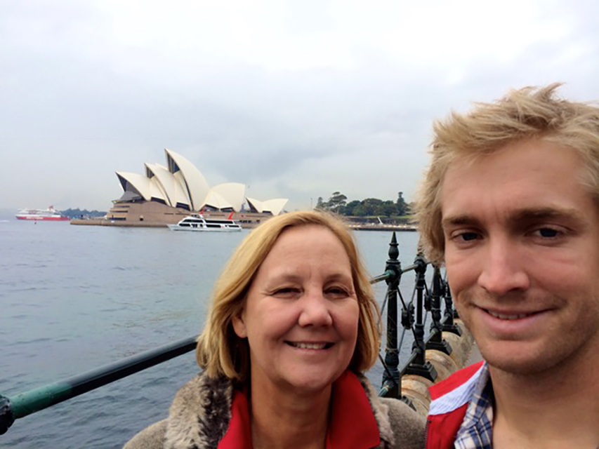 Julia Frey a Caucasian woman in a coat with a fur collar standing in front of Sydney opera house with a young blonde Caucasian man who is smiling.