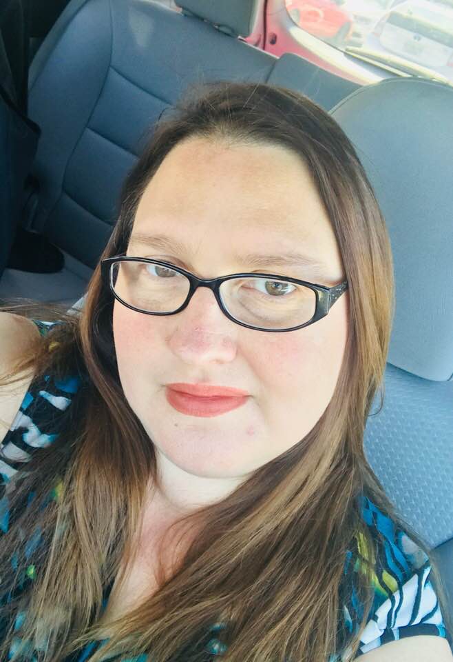 Mandi Clay headshot of a young Caucasian woman in her car. She has long brown hair, glasses and a multi colored top on.