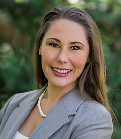 Stephanie Hayes headshot of a smiling Caucasian woman in a grey blazer and white shirt. She is wearing a pearl necklace, has brown eyes and long brown hair. There are trees in the background.