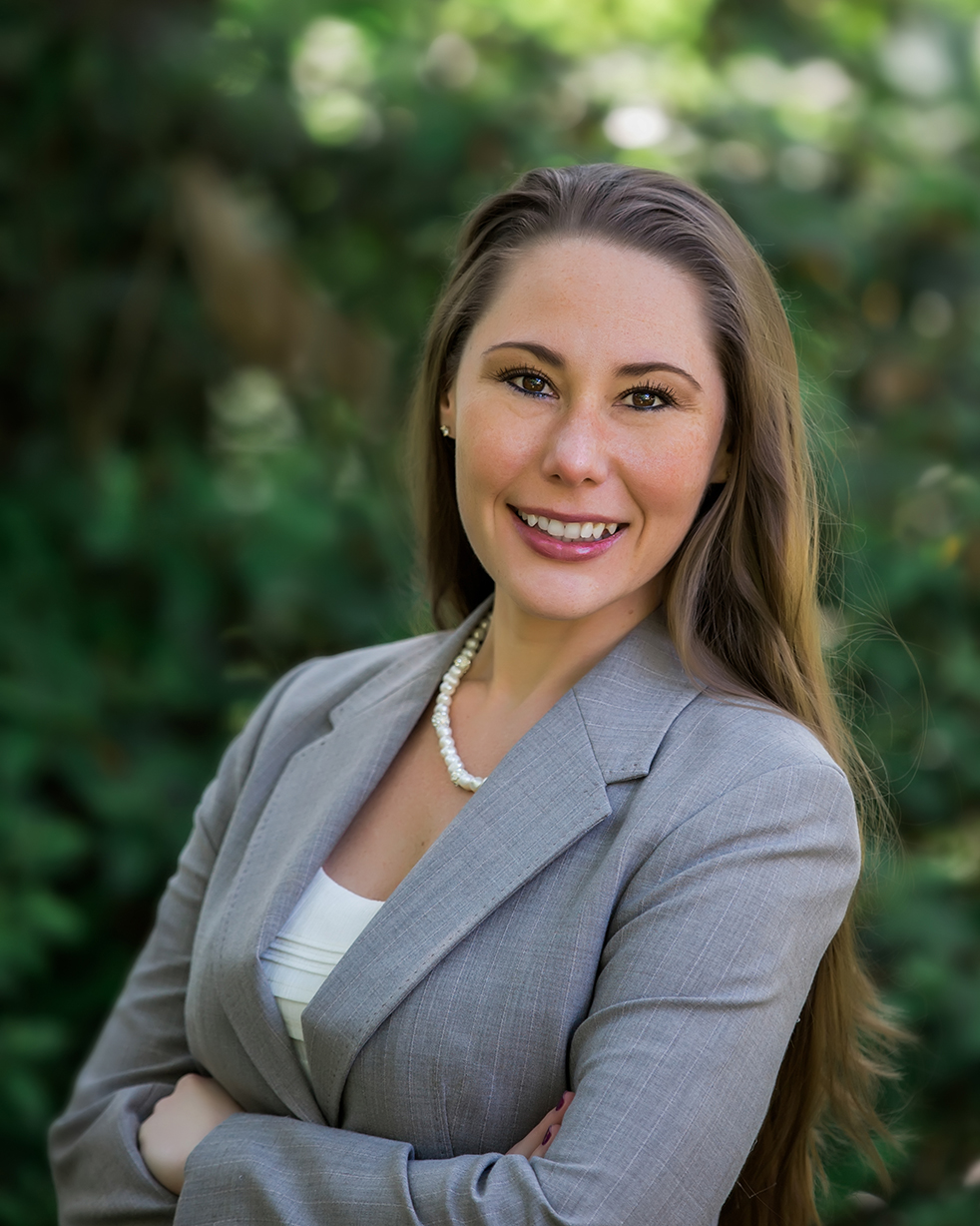 Stephanie Hayes_96dpi Stephanie Hayes headshot of a smiling Caucasian woman in a grey blazer and white shirt. She is wearing a pearl necklace, has brown eyes and long brown hair. There are trees in the background.