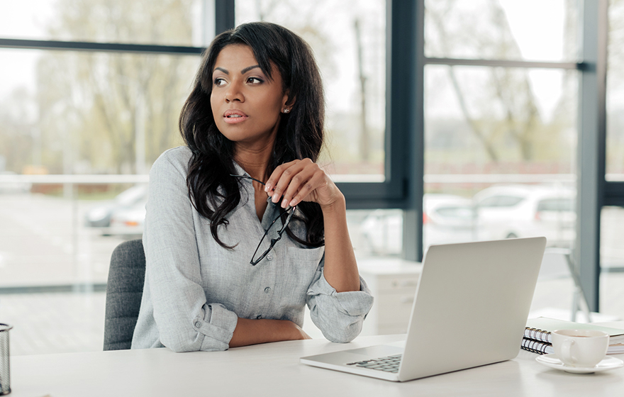 young pensive african american businesswoman in a grey shirt sitting at workplace with a laptop computer, coffee and notebooks