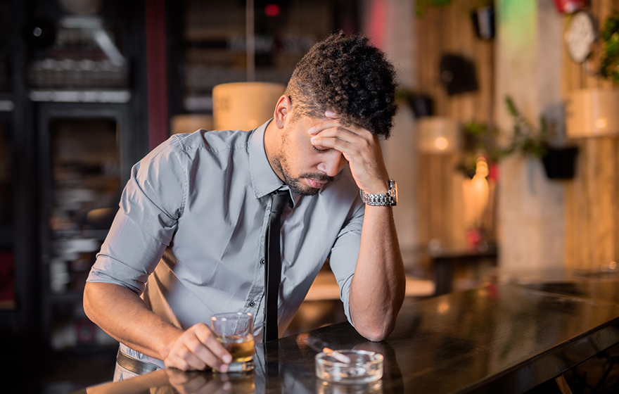 Depressed African American businessman wearing button down shirt and tie in a bar with a drink