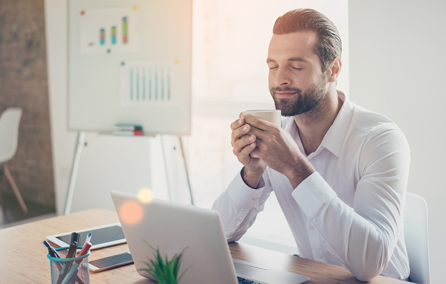 Handsome young Caucasian business man with a beard in white shirt sits at the table in light office and enjoy aroma of coffee