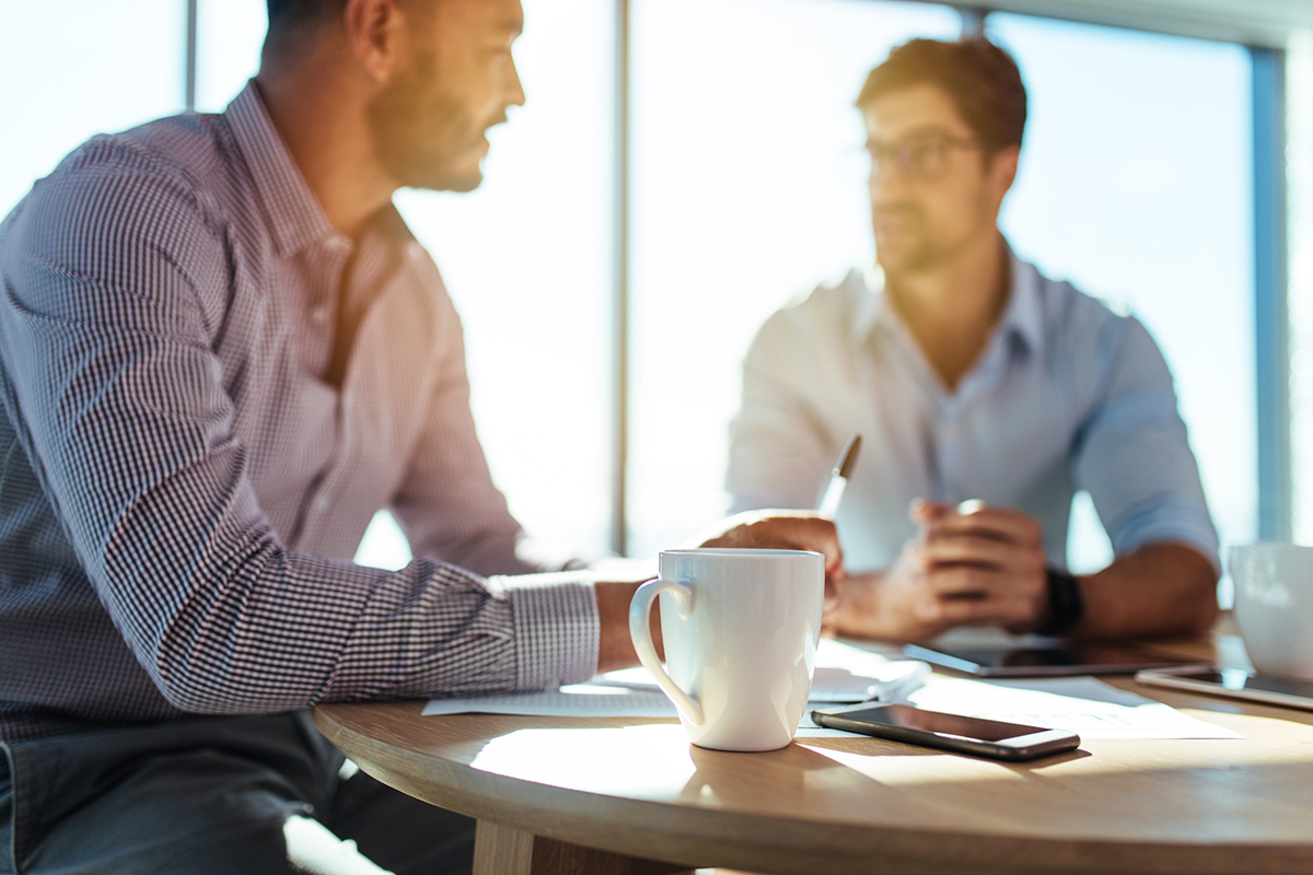 two Caucasian Business men sitting at table in office. They are wearing button down shirts, one has glasses and they both have coffee cups