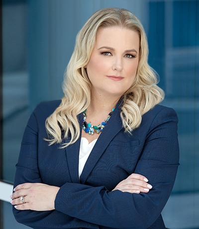 Casseland Cassel a Caucasian woman in a blue blazer with a blue necklace, white shirt and blonde hair headshot on blue background
