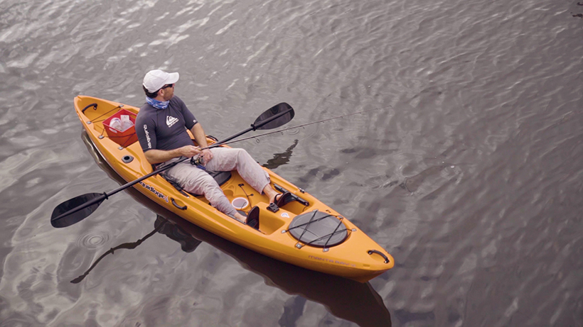 Ron Ponzoli a Caucasian man in a blue shirt, flip flops and a white hat sitting in orange kayak on water