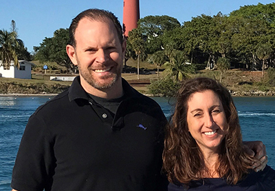 Ron Ponzoli a Caucasian man with glasses, brown hair and a beard and his Caucasian wife with long brown hair. Both are wearing black tops.