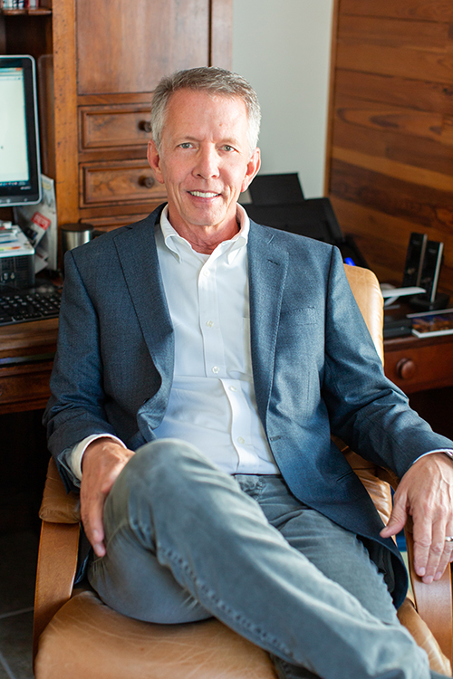 Wayne LaRue Smith an older Caucasian man with a grey sport coat and white shirt and jeans sitting in office chair smiling at camera