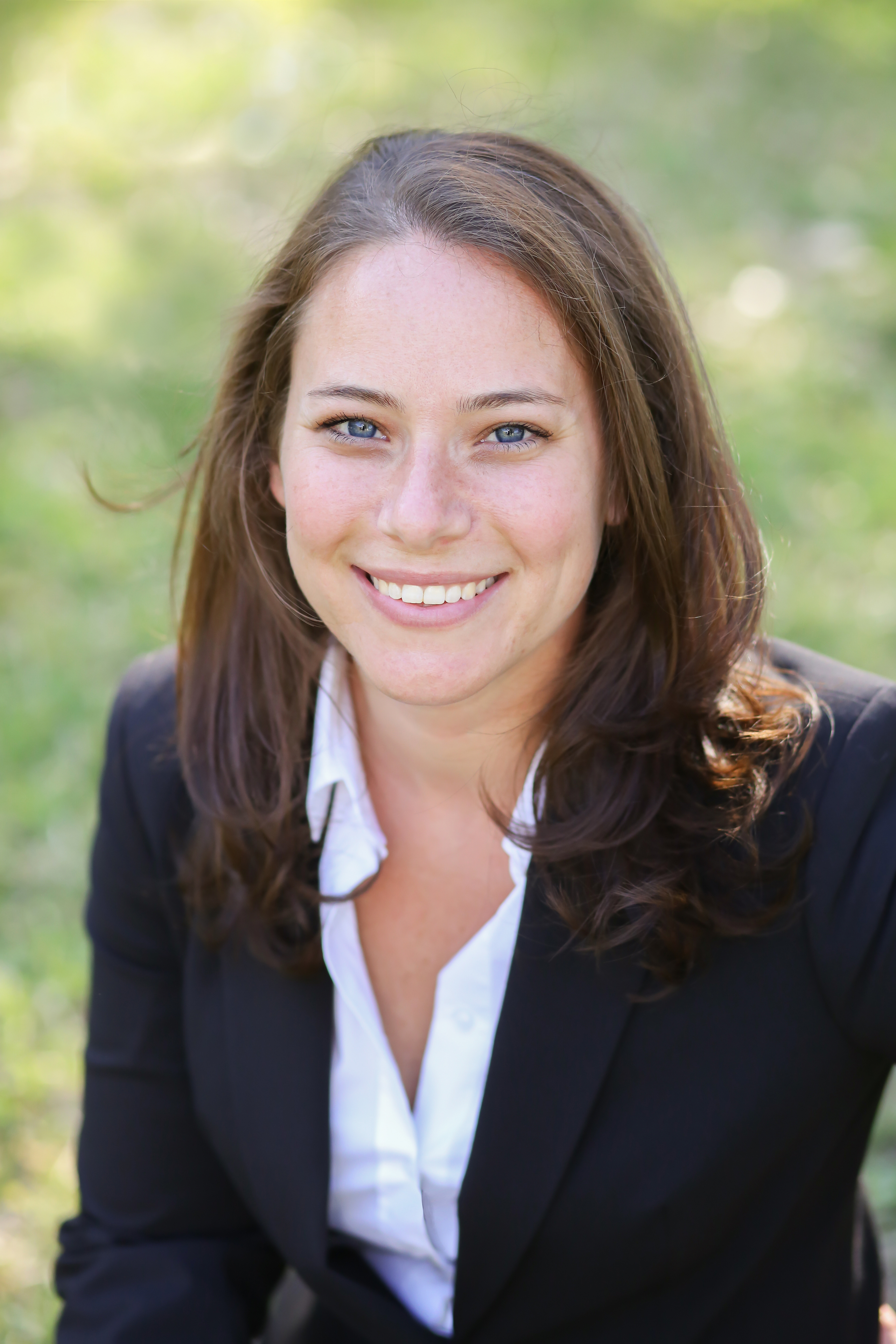 Lifelongstudios-GH-6-Retouched Traci Koster headshot, a young Caucasian woman with blue eyes and long brown hair wearing a white shirt and black blazer with a green grass background
