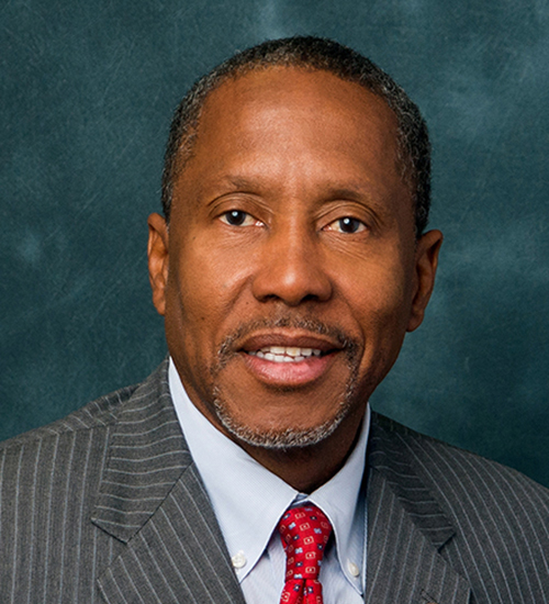 Darryl Rouson headshot, older African American man with brown eyes wearing gray suit and red tie and a gold lapel pin.
