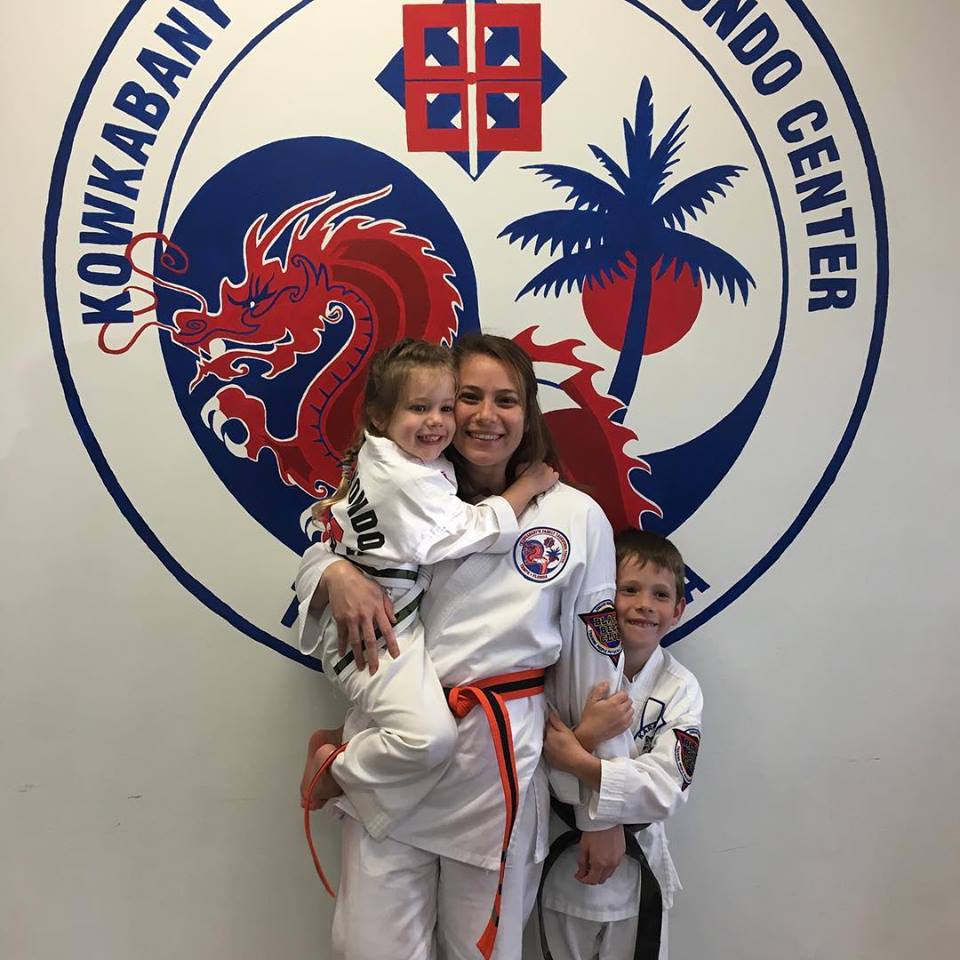 TKD Traci Koster in Taekwondo, a young Caucasian woman with two young Caucasian children, one is a boy and one is a girl, standing behind an Tae Kwon Do background with a dragon and tree symbol