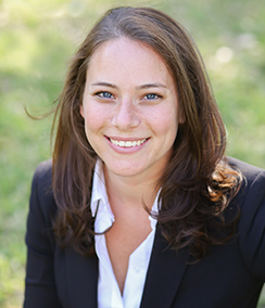 Traci Koster headshot, a young Caucasian woman with blue eyes and long brown hair wearing a white shirt and black blazer with a green grass background