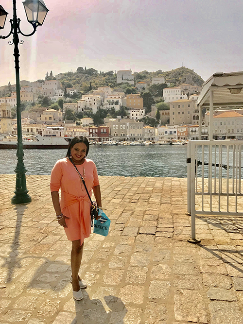 Alison Smith, African American woman in an orange dress and beaded necklace standing in front of historical port city with building on hillside and water/inlet behind her