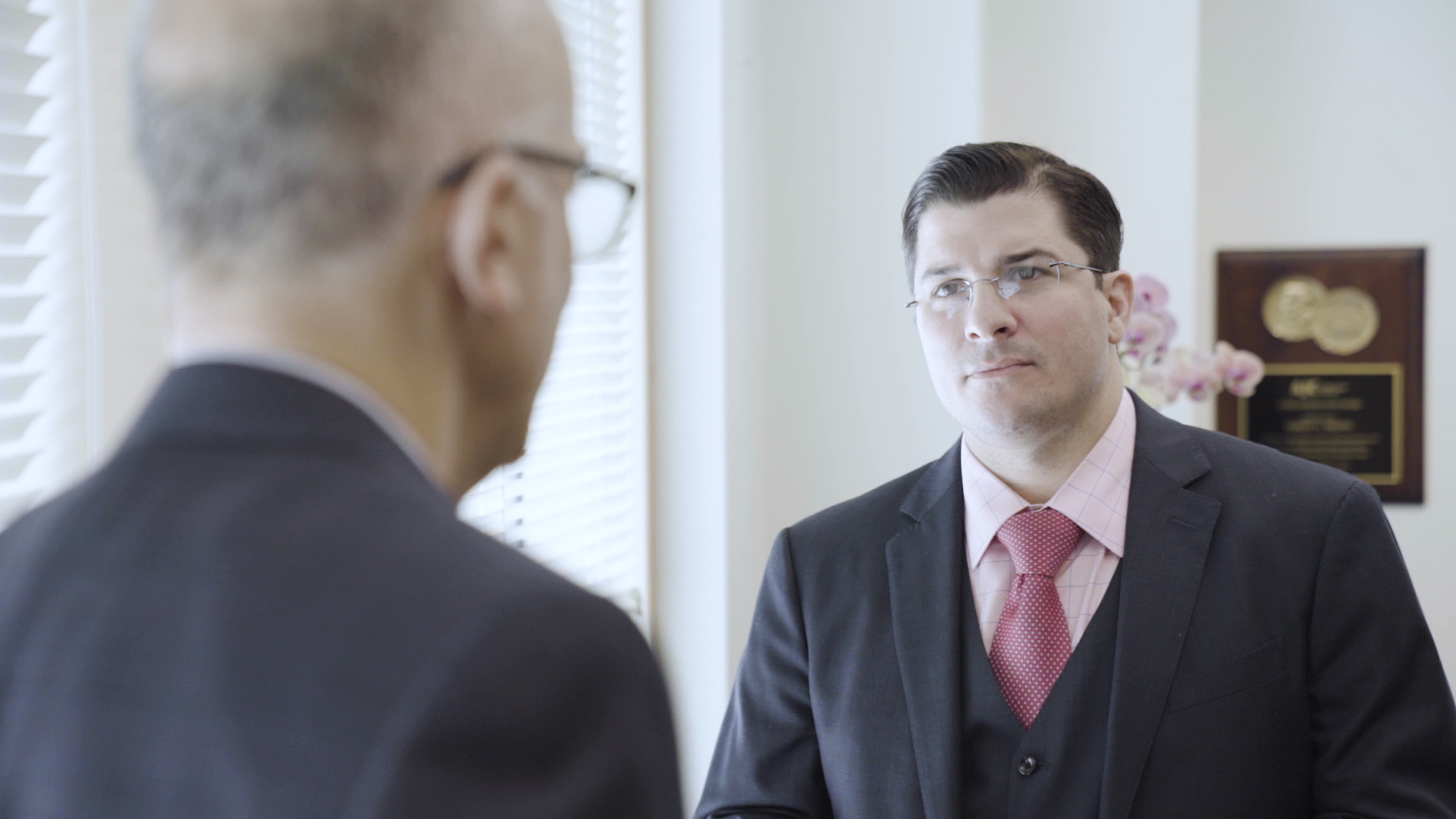 Javier Lopez Latino Man in a grey suit and red tie with an award in the background speaking with an older Caucasian man with his back turned