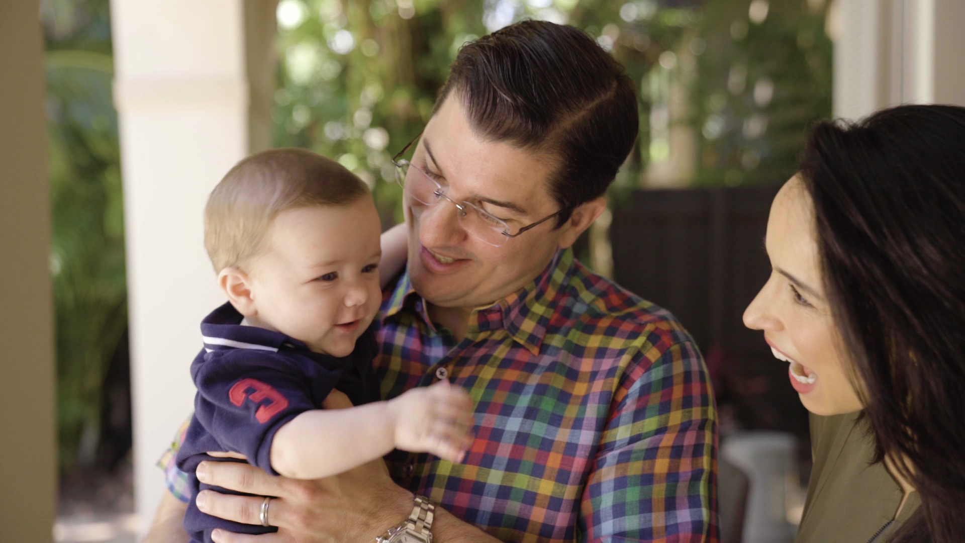 Latino man in glasses and multi colored shirt holding a small infant boy in a blue shirt with a caucasian woman with long brown hair smiling, on a patio outside and sun shining, greenery in background
