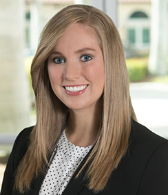 Jennifer Juchnowicz headshot Caucasian woman with blonde in front of window, black blazer, white blouse with black polka dots, long blonde hair, pink lipstick, smiling