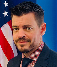 Jeremy Scott headshot, Caucasian man with brown hair, brown eyes and a beard in front of American flag. He is wearing a blue suit, red and blue checkered shirt with a blue tie and a silver tie pin