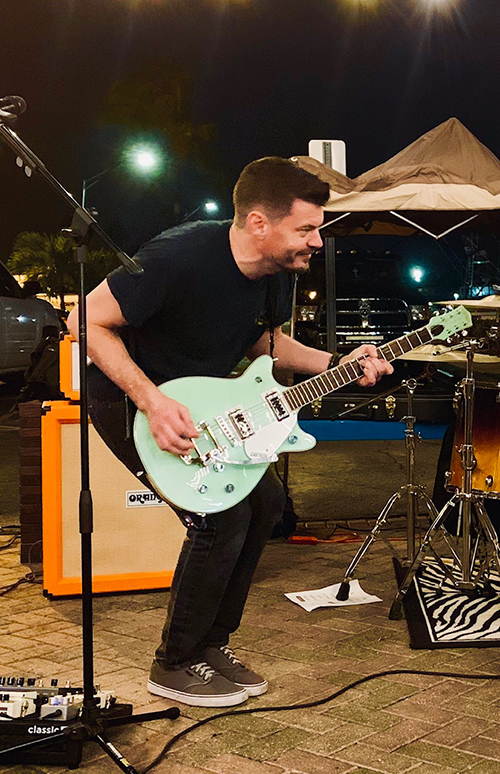 JeremyScott_3 Jeremy Scott a Caucasian man with a beard on stage playing teal electric guitar. Pictured at night with a tent and streetlights in the background