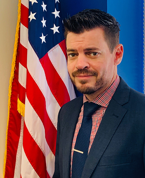 JeremyScott_Edited Jeremy Scott headshot, Caucasian man with brown hair, brown eyes and a beard in front of American flag. He is wearing a blue suit, red and blue checkered shirt with a blue tie and a silver tie pin