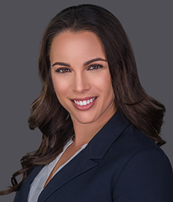 Niki Martell headshot caucasian woman in a dark blue blazer and light blouse with a grey background, long brown hair, brown eyes, smiling