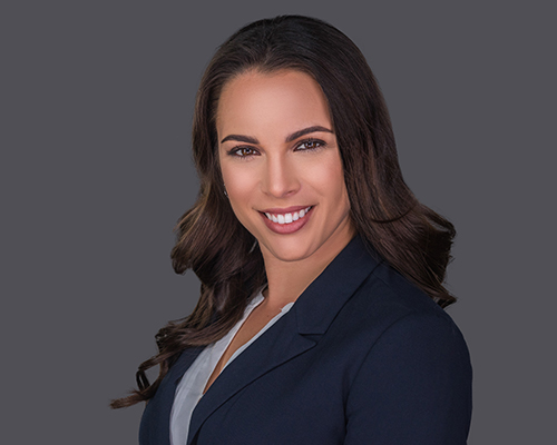 Niki Martell_Edited Niki Martell headshot caucasian woman in a dark blue blazer and light blouse with a grey background, long brown hair, brown eyes, smiling