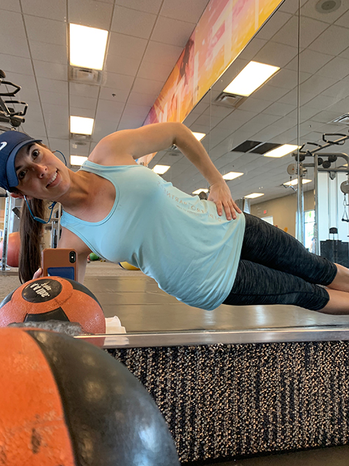 Niki Martell_Workout_Edited Niki Martell, caucasian woman with long black hair in fitness gear and a blue hat performing side plank in a gym environment, mirror in the background, workout equipment and weights