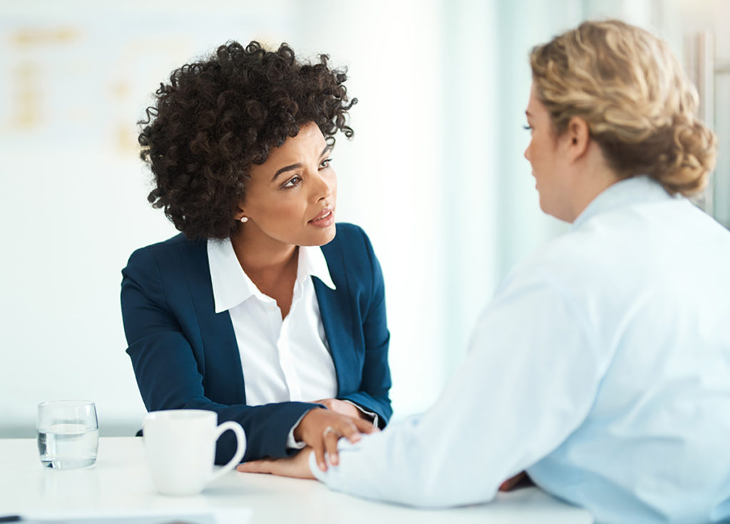 Shot of two businesspeople having a discussion in a modern office, African American woman in a blue sweater with white shirt and pearl earrings and curly hair comforting a caucasian woman with blonde curly hair in a bun wearing a light colored shirt