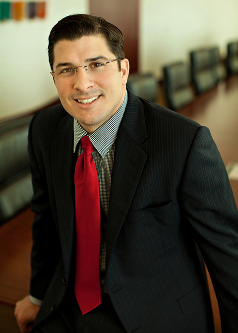 Javier Lopez Latino Man in a grey suit and red tie and glasses sitting in front of long table framed by black chairs