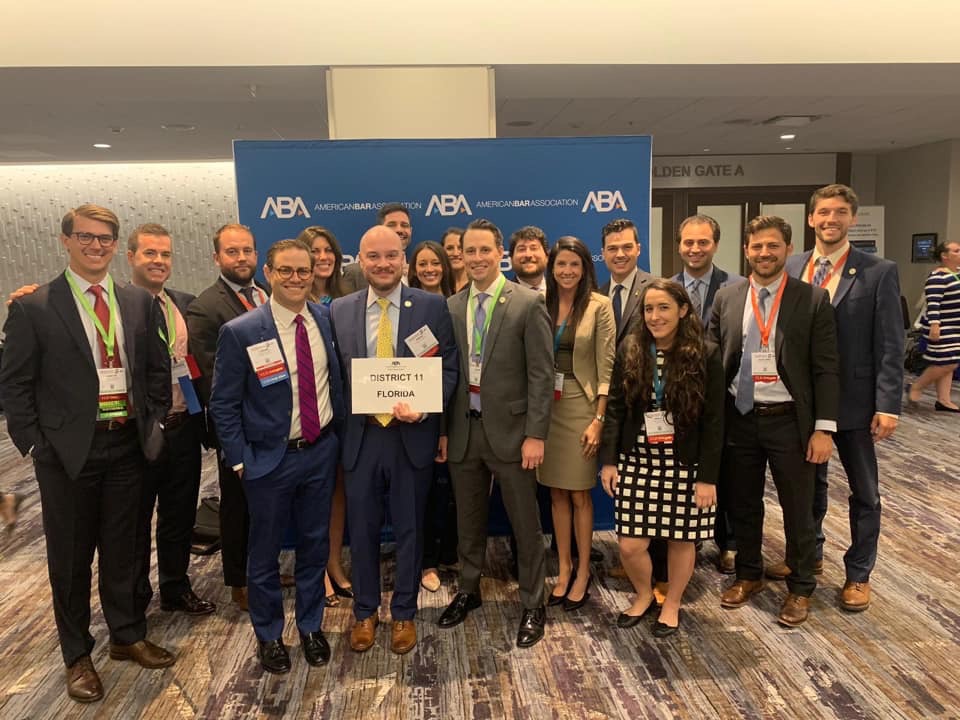 American Bar Association Annual Convention Report Meeting Image, twenty professional caucasian people standing and smiling, business suits, dresses, blazer, American Bar Association Logo on Blue Banner in Background
