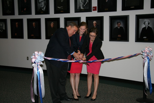 Lactation Room 3 Ribbon cutting, red and blue ribbon, at courthouse, two caucasian women in red dresses with black blazers, caucasian man in a black business suit, professional dress