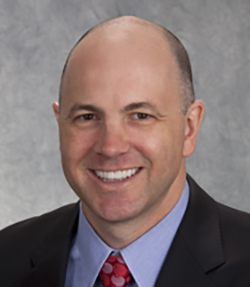 Damian C. Taylor headshot of a middle aged smiling Caucasian man wearing a black suit with a blue shirt and red tie.