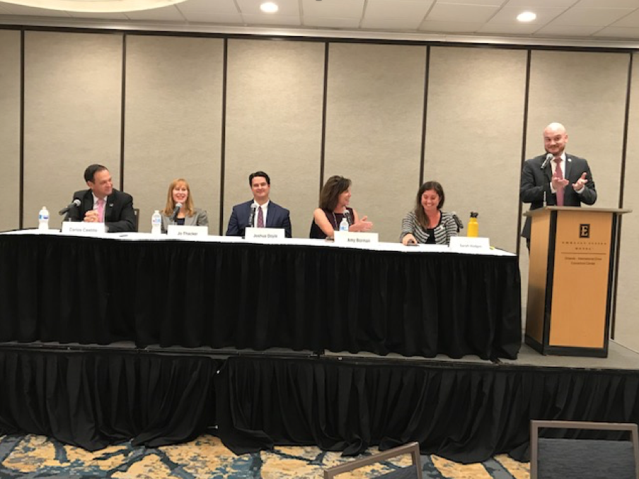 Government Lawyers Committee Update 2 man clapping behind podium, five people seated at a table, six Caucasian people, business suits, blazers, professional dress